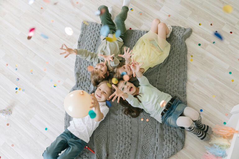 Aerial view of happy children celebrating with balloons and confetti indoors on a rug.